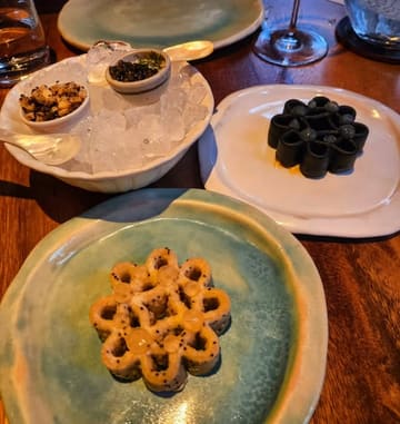 A high-quality, top-down shot of a fine dining tasting menu course at a Michelin-starred Thai restaurant. The image features intricate, flower-shaped crackers served with caviar and other accompaniments, presented on artisanal ceramic plates.