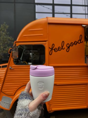 A casual, lifestyle-focused shot featuring a reusable coffee cup held in front of a branded orange food truck. The image captures a moment of post-workout refreshment, effectively using the branded vehicle as a backdrop to establish the venue's identity.