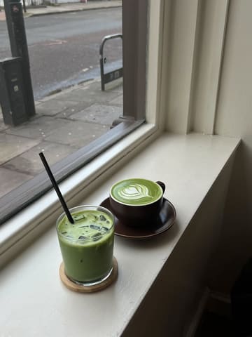 A simple, candid shot of two matcha drinks—one hot and one iced—placed on a windowsill, capturing a quiet moment in a cafe setting.