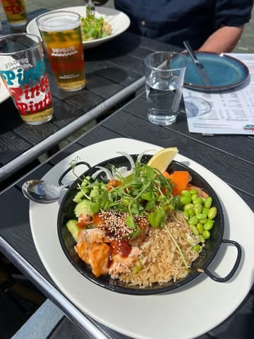 A casual, outdoor shot of a teriyaki salmon poke bowl served in a metal pan, accompanied by a beer and water glass on a black table.