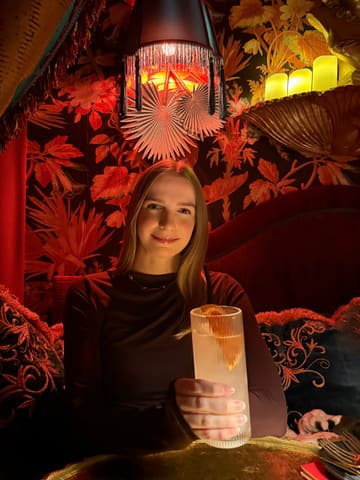 A portrait of a woman holding a cocktail in a dimly lit, ornately decorated bar, showcasing the venue's intimate and stylish atmosphere.