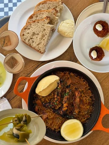 A high-quality, overhead flat-lay shot of a Spanish-inspired dinner spread at a restaurant. The image features a central pan of prawn fideuà, accompanied by bread, croquetas, and gilda skewers, all arranged neatly on a rustic wooden table. The lighting is bright and natural, highlighting the textures of the food effectively.