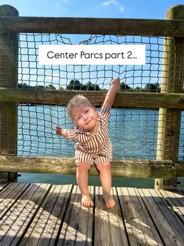 A bright, high-quality lifestyle shot of a toddler sitting on a wooden bridge over a lake at a Center Parcs holiday resort, capturing a family-friendly travel moment.