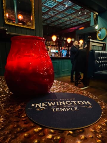 A close-up shot of a table in a pub, featuring a red glass candle holder and a branded coaster for 'Newington Temple'. The background shows a warm, dimly lit pub interior with patrons.