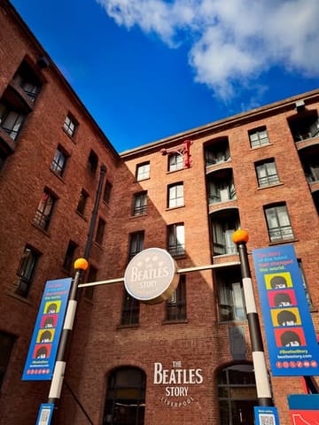 A low-angle, wide shot of The Beatles Story museum entrance at the Albert Dock in Liverpool, capturing the iconic red brick architecture and signage against a bright blue sky.