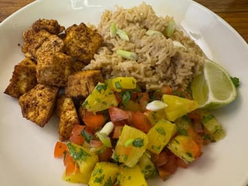 A top-down, functional shot of a home-cooked meal featuring jerk tofu, coconut rice, and pineapple salsa. The image captures the components clearly but lacks the professional lighting and deliberate plating required for high-end hospitality marketing.