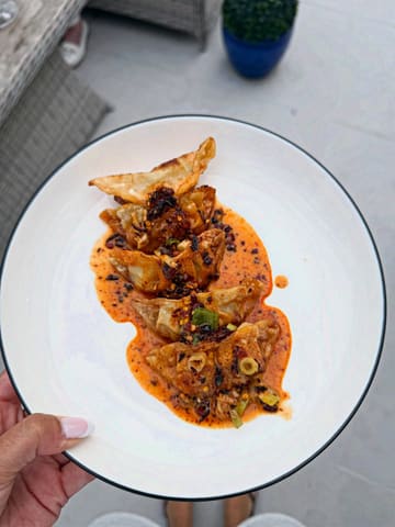 A high-quality, overhead shot of a plate of pan-fried dumplings drizzled with chili oil, held by a person outdoors. The lighting is natural and even, highlighting the texture of the dumplings and the vibrant color of the sauce.