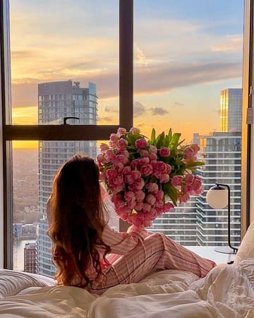 A high-quality, aspirational lifestyle shot featuring a person sitting on a bed in a hotel room, holding a large bouquet of pink tulips against a backdrop of a city skyline at sunset. The image effectively communicates a romantic or celebratory atmosphere.