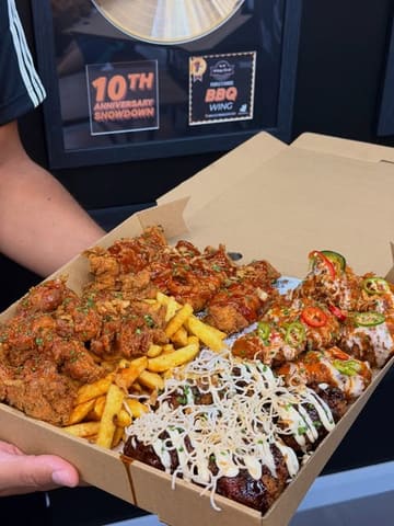 A first-person, eye-level shot of a large, overflowing 'Feast Box' containing various styles of chicken wings, tenders, and loaded fries, held up against a wall displaying award certificates.