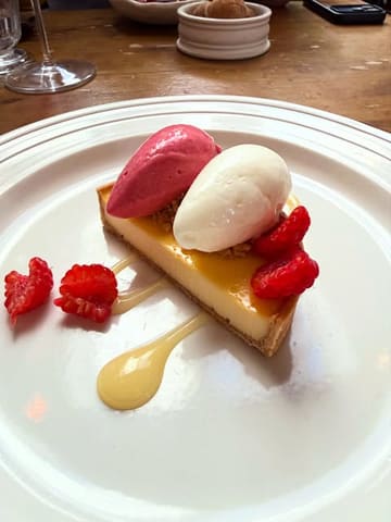 A high-quality, close-up shot of a raspberry custard tart served with two quenelles of sorbet and cream, garnished with fresh raspberries and a sauce drizzle, presented on a white plate at a restaurant.
