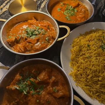 A top-down, overhead shot of three metal bowls filled with various curries and a large plate of yellow rice, arranged on a dark marble table. The image highlights the textures and vibrant colours of the food, presenting a classic Indian dining experience.