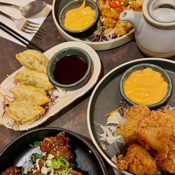 A flat-lay photograph of a spread of Japanese-inspired dishes, including gyoza, karaage, and various noodle and rice dishes, arranged on a dark table.
