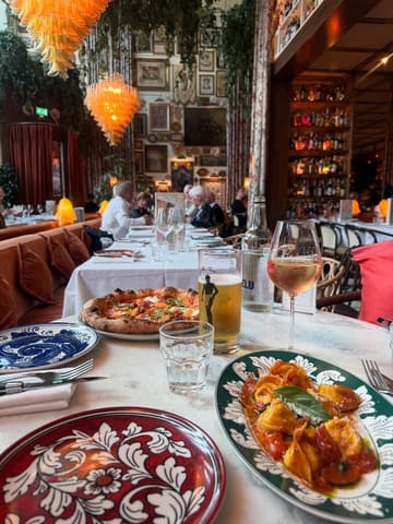 A high-quality, inviting shot of a table at La Bellezza, featuring a pizza and a pasta dish, set against the restaurant's signature maximalist, ornate interior. The warm lighting and well-composed angle create an appealing atmosphere that highlights the dining experience.