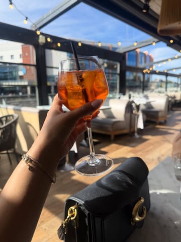 A bright, inviting POV shot of an Aperol Spritz held up against a sunlit, outdoor terrace setting, effectively capturing a summer atmosphere.