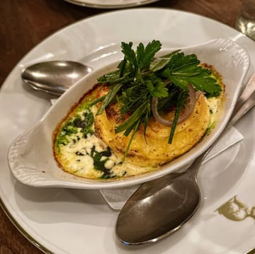 A close-up, eye-level shot of a cheese soufflé served in an oval dish, garnished with fresh herbs and red onion, presented on a white plate with a spoon.