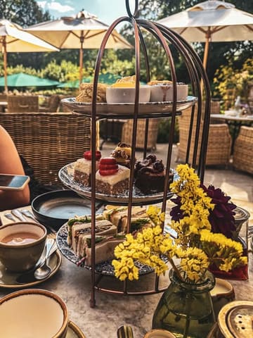A high-angle shot of an afternoon tea service presented on a birdcage-style stand, set on a table in an outdoor garden. The spread includes sandwiches, cakes, and scones, accompanied by a cup of tea and a vase of yellow flowers.