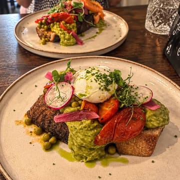 A close-up, high-angle shot of a brunch dish featuring thick sourdough toast topped with smashed avocado, a poached egg, roasted tomatoes, and microgreens, served at a cafe.