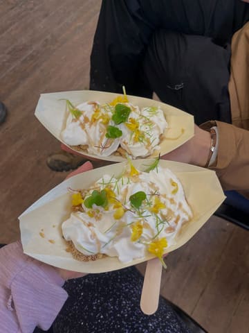 A top-down shot of two people holding small wooden boats containing a dessert garnished with edible flowers and herbs, captured during an outdoor foraging experience.