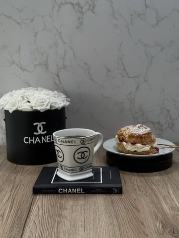 A staged lifestyle shot featuring a cream scone and a cup of coffee, heavily accessorized with Chanel-branded items including a flower box and a book, set against a marble-effect background.