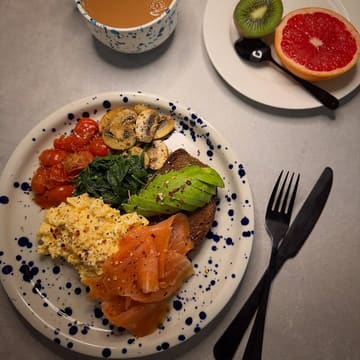 A well-composed, overhead photograph of a healthy brunch plate featuring scrambled eggs, smoked salmon, avocado toast, sautéed mushrooms, spinach, and roasted tomatoes, served with a side of grapefruit and kiwi. The image is clean, bright, and visually appealing, showcasing a balanced meal.