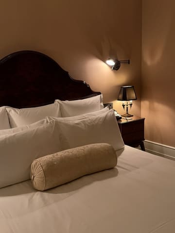 A moody, atmospheric shot of a hotel bed at The Ned London, focusing on the warm, inviting lighting and clean, well-made bedding. The image captures a sense of luxury and comfort, typical of a high-end hotel stay.