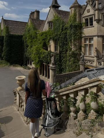 A high-quality, aesthetic shot of a visitor walking up the stone steps towards a historic, ivy-clad manor house in the Cotswolds, carrying a golf bag. The image captures a serene, 'old money' countryside atmosphere, emphasizing the luxury and leisure aspects of the venue.