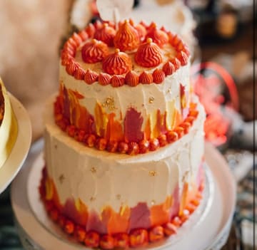 A vibrant, two-tier wedding cake featuring an abstract, textured frosting design in shades of orange, pink, and white, accented with gold leaf and piped rosettes. The cake is presented on a stand, though the composition suffers from tight framing that clips the top of the cake topper.