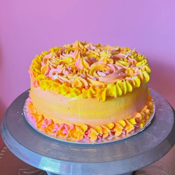 A vibrant, multi-colored vanilla sponge cake with strawberry jam filling, displayed on a metal cake turntable against a plain pink background.