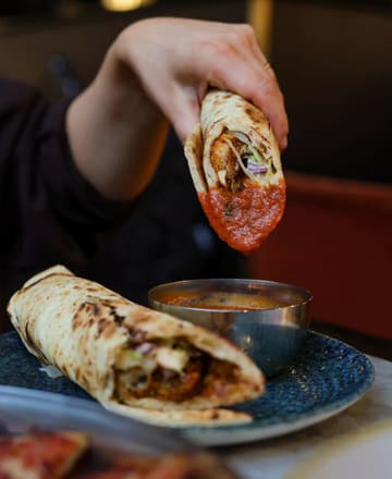 A close-up, high-contrast shot of a chicken pakora naan wrap being dipped into a vibrant red sauce, with a second wrap visible in the foreground. The lighting is dramatic, focusing attention on the texture and sauce of the wrap.