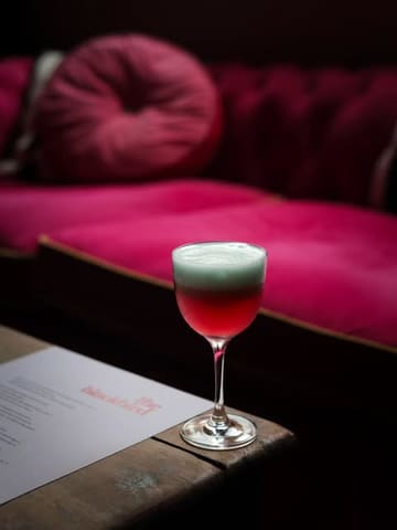 A moody, atmospheric shot of a cocktail in a coupe glass, set against a backdrop of pink velvet seating at The Blackbird in Edinburgh. The lighting is low and intimate, highlighting the drink's foam while keeping the venue's interior soft and out of focus.