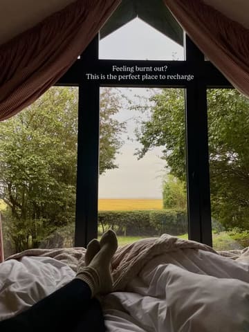 A serene, first-person perspective shot from inside a cabin, looking out through a large floor-to-ceiling window at a nature reserve. The foreground features a pair of feet in socks resting on a bed, creating an immediate sense of relaxation and comfort. The text overlay reinforces the theme of recharging and escaping burnout, perfectly aligning with the tranquil visual.