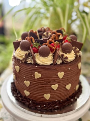 A high-quality, close-up photograph of a decadent, multi-layered chocolate cake decorated with various toppings including chocolate truffles, pretzels, strawberries, and white chocolate hearts. The cake is presented on a white cake stand, with a soft-focus background.