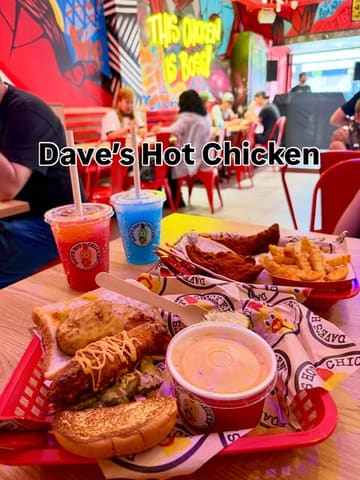 A casual, honest shot of a meal at Dave's Hot Chicken, featuring their signature fried chicken, fries, mac and cheese, and colourful drinks. The image captures the vibrant, graffiti-filled interior of the venue, though the lighting and busy background detract from the food itself.