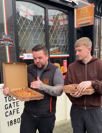 Two men stand outside a takeaway shop holding pizza boxes, likely filming a review or recommendation video for local food spots in the Bury and Bolton area.