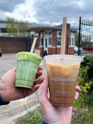 A first-person perspective shot holding two iced drinks, a matcha latte and an iced coffee, in front of a cafe building. The image captures the drinks clearly against the venue's exterior.