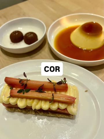 A top-down, overhead shot of three dessert plates arranged on a wooden table, featuring a rhubarb dessert, a flan, and two chocolate truffles at a restaurant called COR.