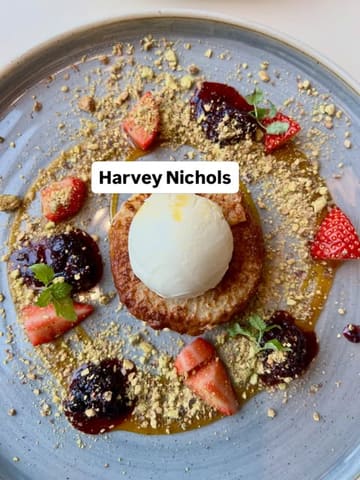 A high-quality, top-down photograph of a plated Pain Perdu dessert served at Harvey Nichols. The dish is elegantly presented with a scoop of ice cream, fresh strawberries, a berry compote, and a dusting of crushed pistachios on a grey plate, set against a clean, neutral background.