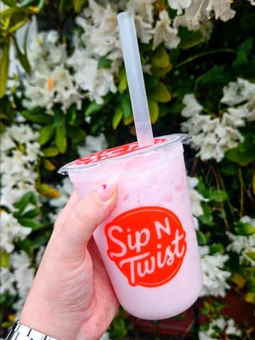 A close-up, eye-level shot of a hand holding a pink fizzy drink in a branded plastic cup, set against a backdrop of white flowers.