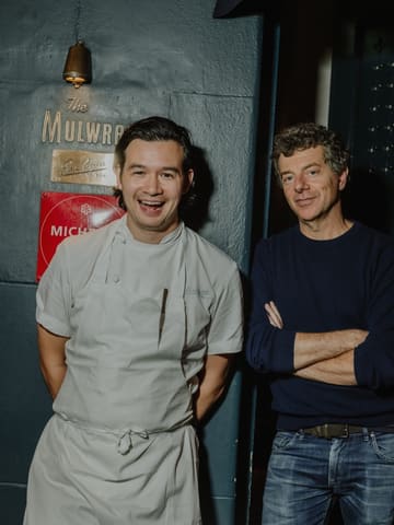 A portrait of a chef and another individual standing in front of a wall featuring a Michelin plaque, likely taken at Evelyn's Table. The image captures a candid, happy moment between the subjects.
