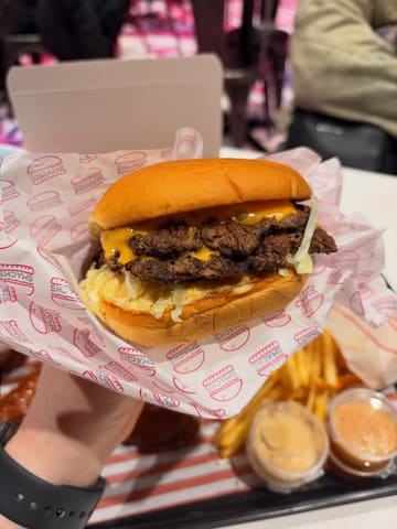 A close-up, handheld shot of a double-patty smash burger in a branded wrapper, with fries and dipping sauces visible on a tray in the background.