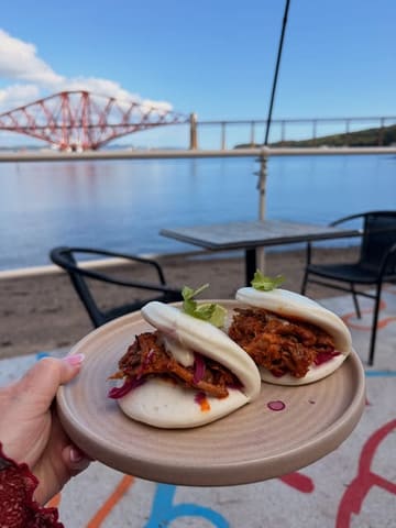 A high-quality, well-composed shot featuring BBQ pork bao buns held in the foreground against the iconic backdrop of the Forth Bridge in South Queensferry. The natural lighting and clear focus on the food make for an appealing and shareable image.