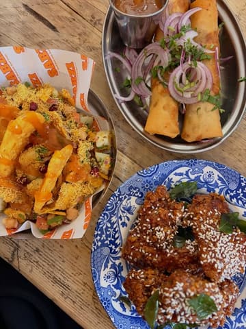 A high-quality, overhead flat-lay photograph of three distinct dishes arranged on a rustic wooden table. The composition is clean and balanced, showcasing a variety of textures, colors, and garnishes, including spring rolls, sesame-crusted fried chicken, and a vibrant chaat dish.