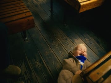 A candid, atmospheric lifestyle shot capturing a joyful moment of a child lying on a rustic wooden floor, likely inside a holiday rental cabin in Wales. The lighting is moody and warm, emphasizing the cozy, off-grid nature of the stay.
