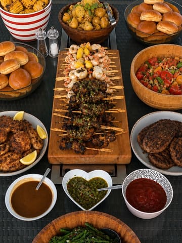A professional, overhead flat-lay photograph of a large BBQ catering spread, featuring skewers, burgers, corn, and various salads, arranged neatly on a dark outdoor table.
