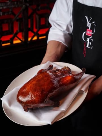 A professional, high-quality promotional shot featuring a chef presenting a whole roasted duck at Yú Gé Cantonese Cuisine. The lighting and composition highlight the texture and presentation of the dish, conveying a premium dining experience.