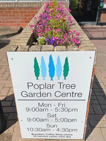 A clear, well-lit photograph of a sign for Poplar Tree Garden Centre, set against a backdrop of flowers. The image provides essential information about the venue, including opening hours, and establishes the location's identity.