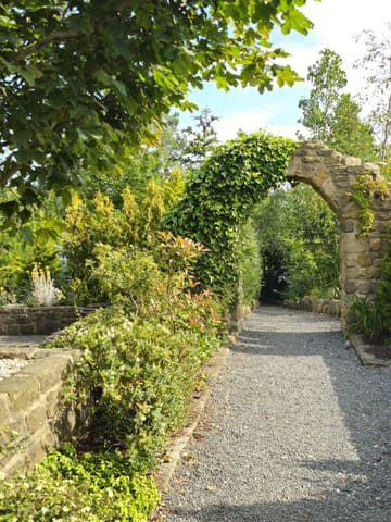 A scenic, eye-level shot of a gravel path leading through a stone archway covered in ivy at a garden centre, surrounded by lush greenery and trees under a bright, sunny sky.