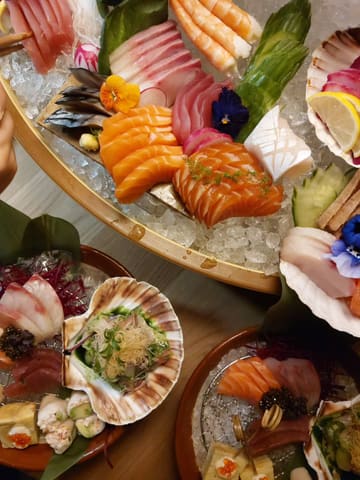 A top-down, high-angle shot of a Japanese sushi and sashimi platter served on ice, likely at a restaurant launch event. The image captures a variety of fresh fish, including salmon and tuna, arranged on a wooden boat-style platter, alongside other smaller dishes in the foreground.