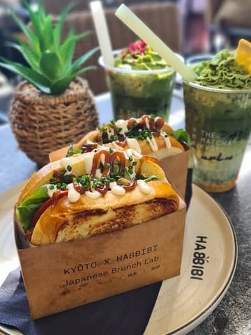 A bright, well-composed close-up shot of two Japanese-style pocket sandwiches in branded paper packaging, served alongside two matcha drinks on a table, likely at a rooftop venue.
