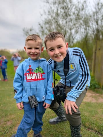 A high-quality, candid photograph of two young boys smiling at the camera while outdoors, wearing binoculars, suggesting a nature-based activity or exploration experience.